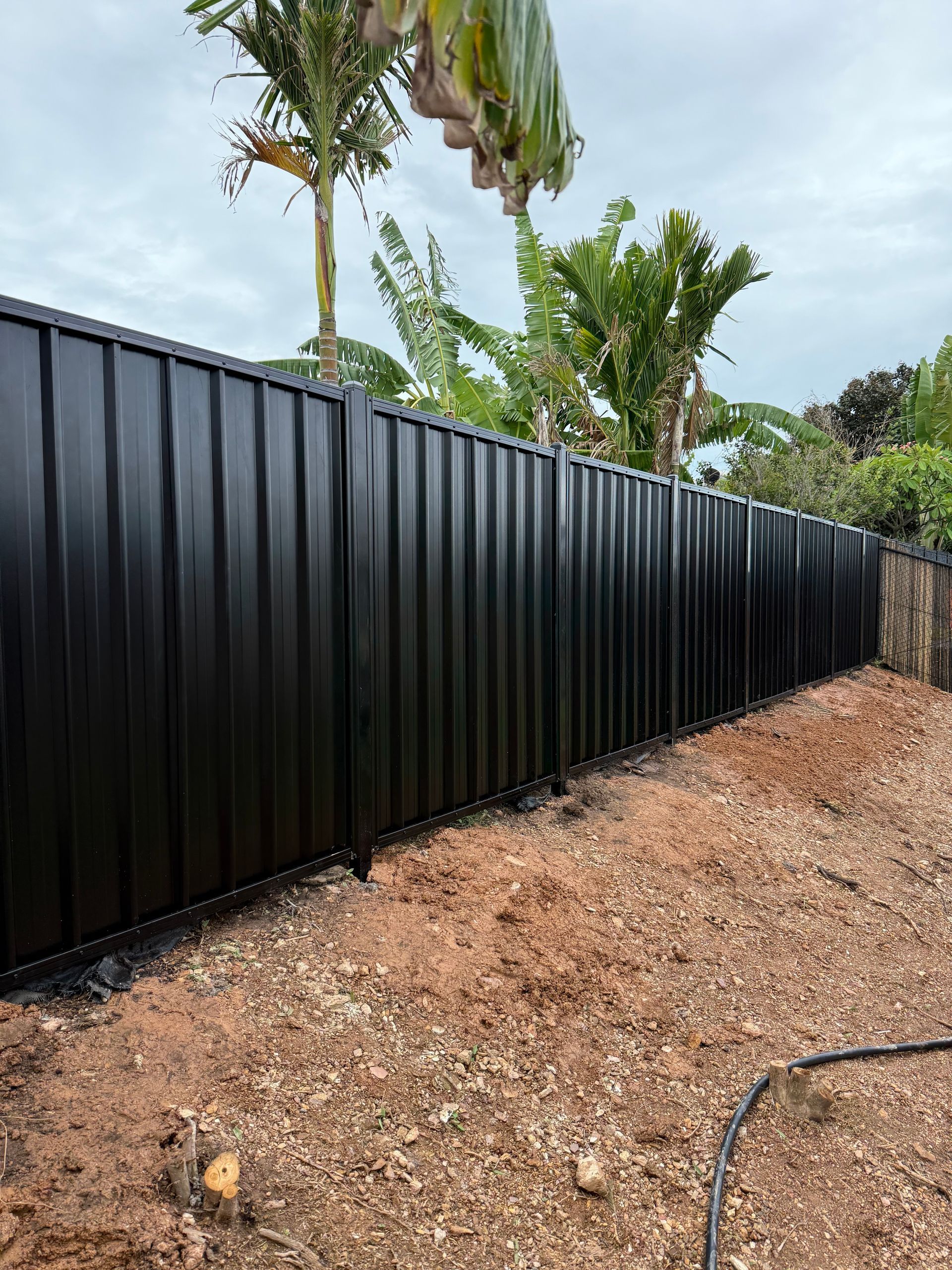 Black corrugated metal fence in a yard with dirt and plants in the background. — True North Fencing In Noonamah, NT