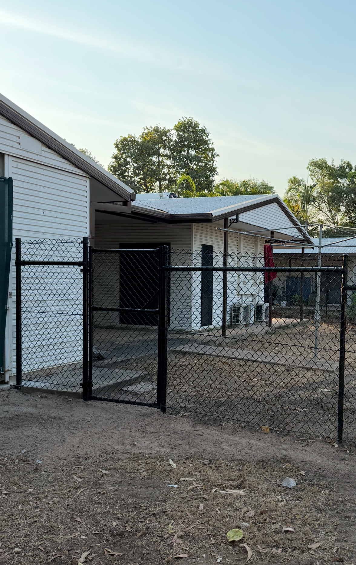 Black chain-link fence surrounds a white building with trees in the background under a blue sky. — True North Fencing in Noonamah, NT