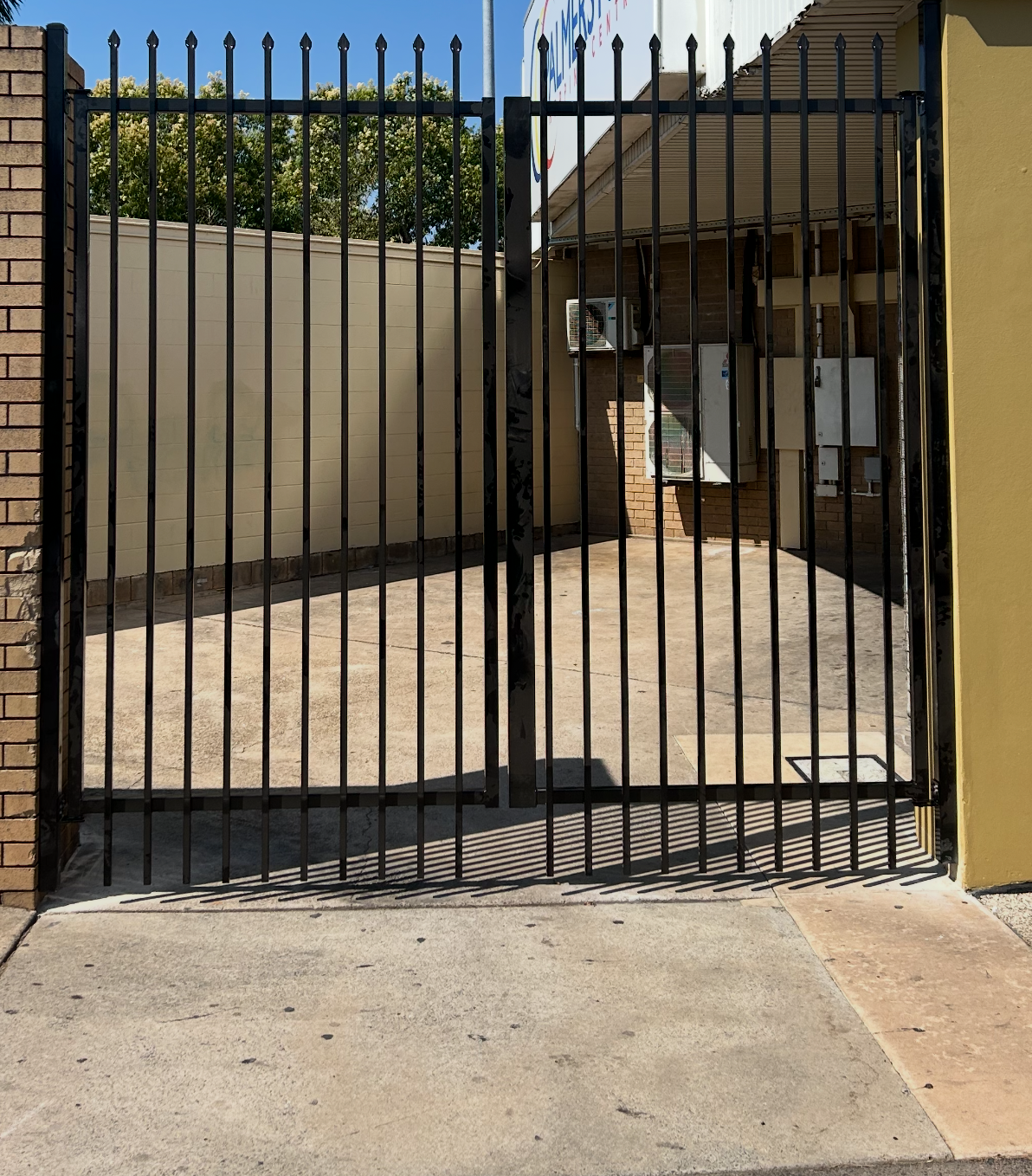 Black metal gate in a concrete driveway, between brick and yellow walls. — True North Fencing In Noonamah, NT