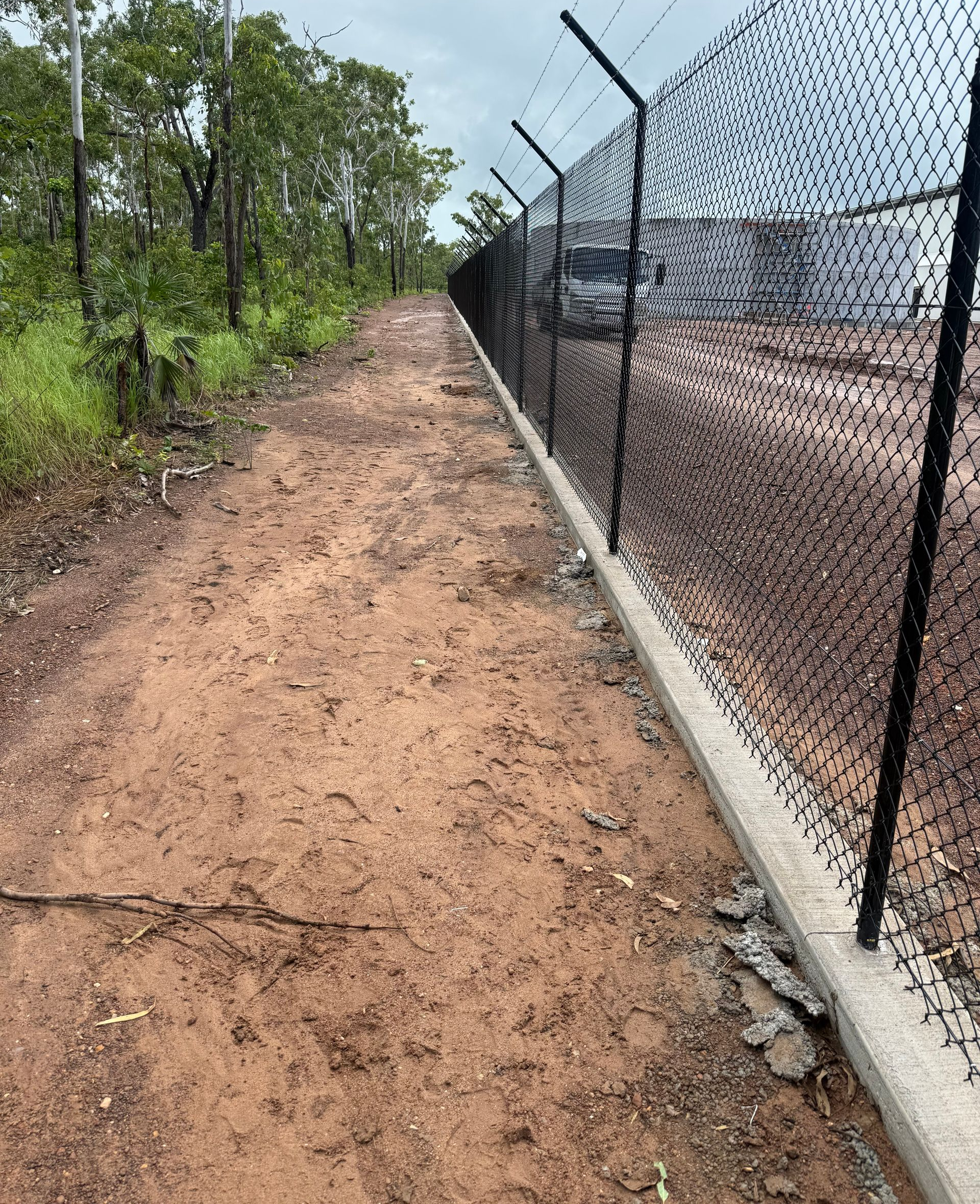 Dirt path beside a black chain-link fence topped with barbed wire, leading toward a building. — True North Fencing In Noonamah, NT