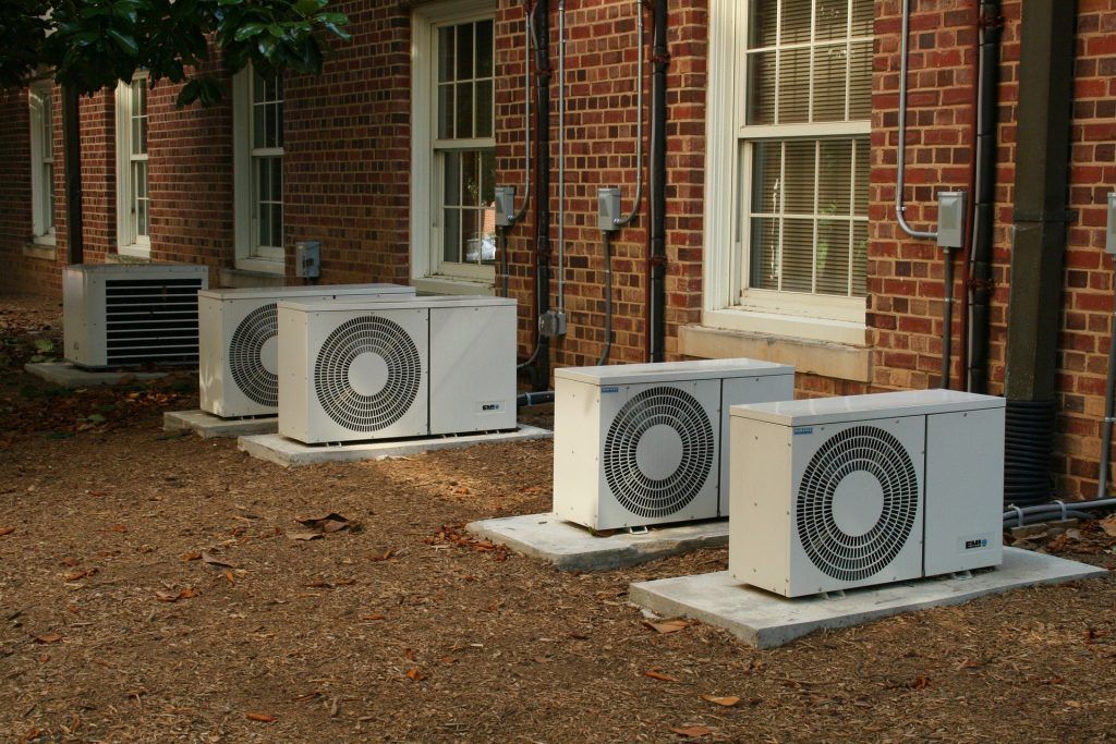Air conditioning units lined up against a brick building with windows. Gravel ground.