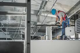 A construction worker in a blue hard hat and safety harness works on pipes and ductwork on a metal ceiling platform.