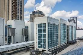 Row of large rooftop HVAC units and ducts on a city building under a partly cloudy sky