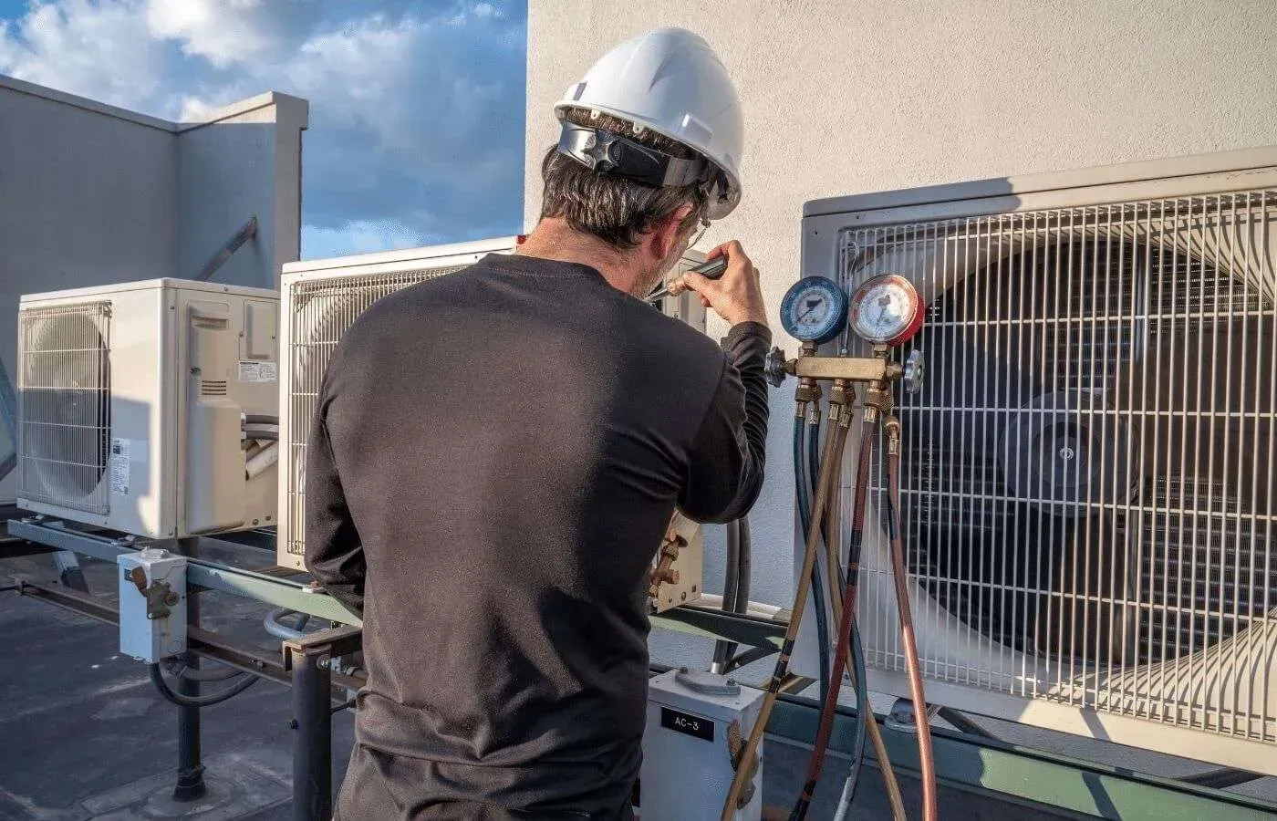 Technician in hard hat inspecting rooftop commercial HVAC units with gauges and hoses
