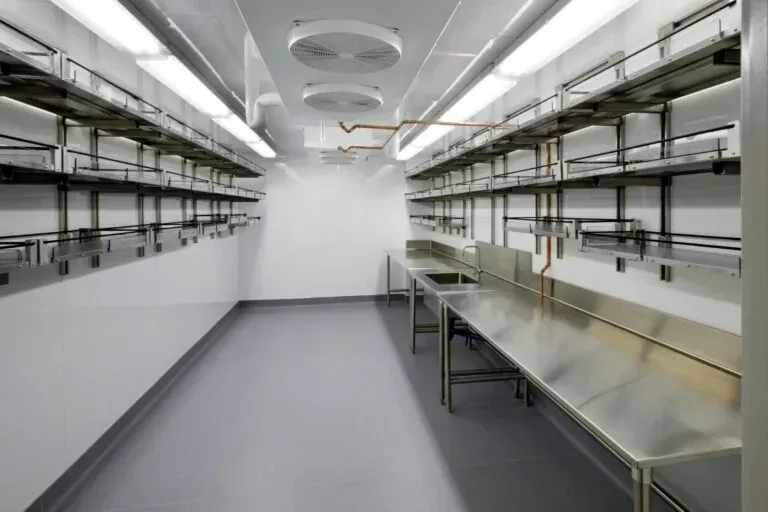 Empty commercial fridge with stainless steel counters and shelves, white walls, and gray floor