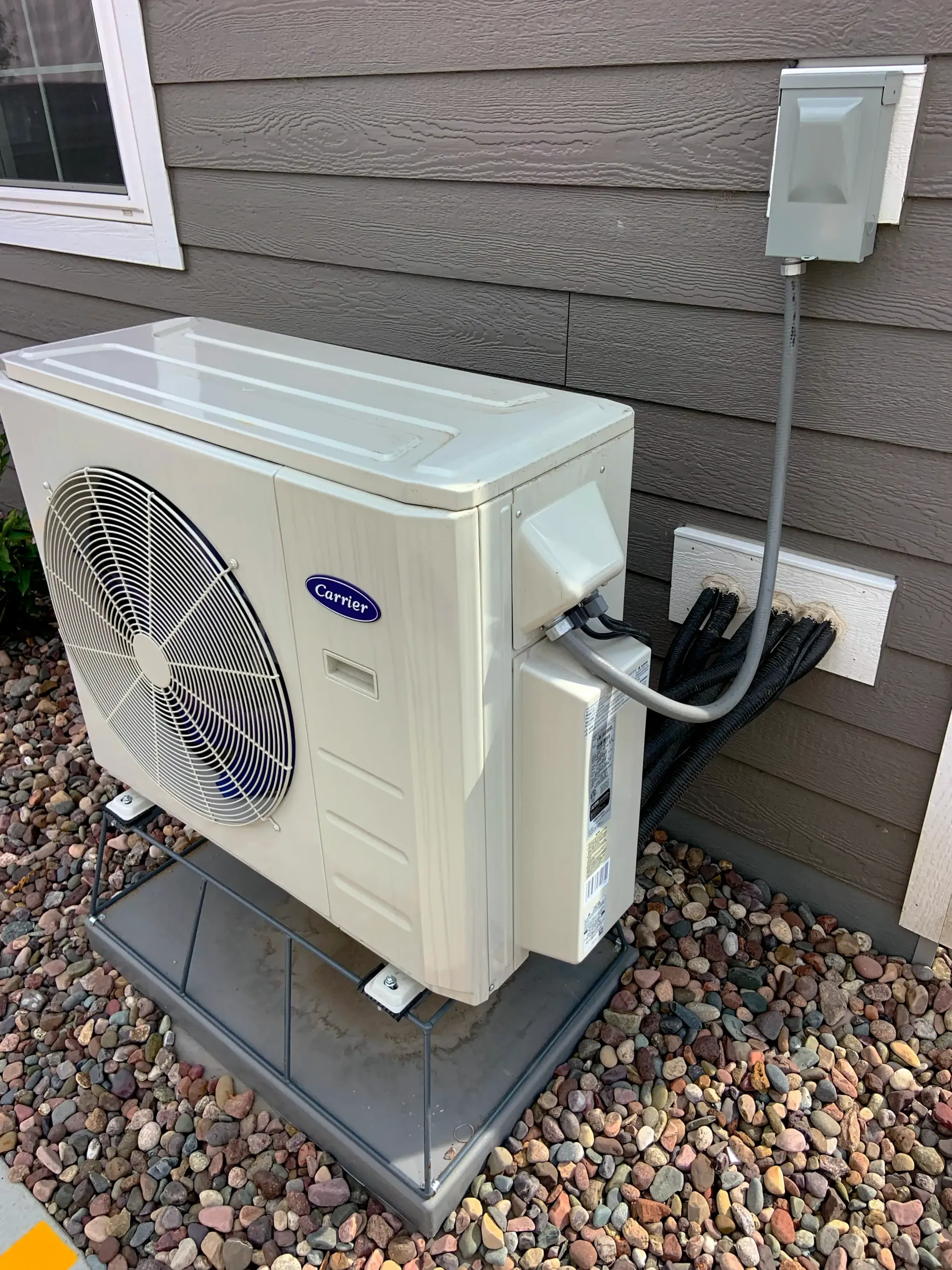 Carrier heat pump unit mounted on a grey pad against a grey house exterior
