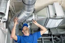 A technician in a blue shirt inspecting metal HVAC ductwork installed on a ceiling.