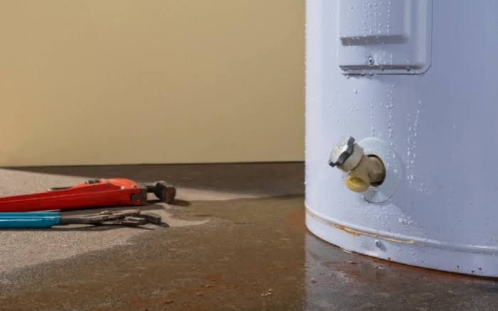 Red pliers on a dusty floor beside a light blue hot water tank with a valve