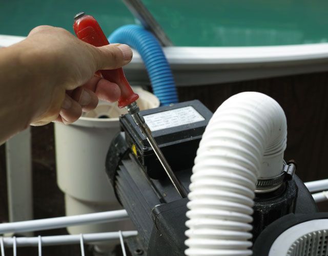 Hand with red screwdriver tightening a screw on a pool pump, near pool edge and hoses.