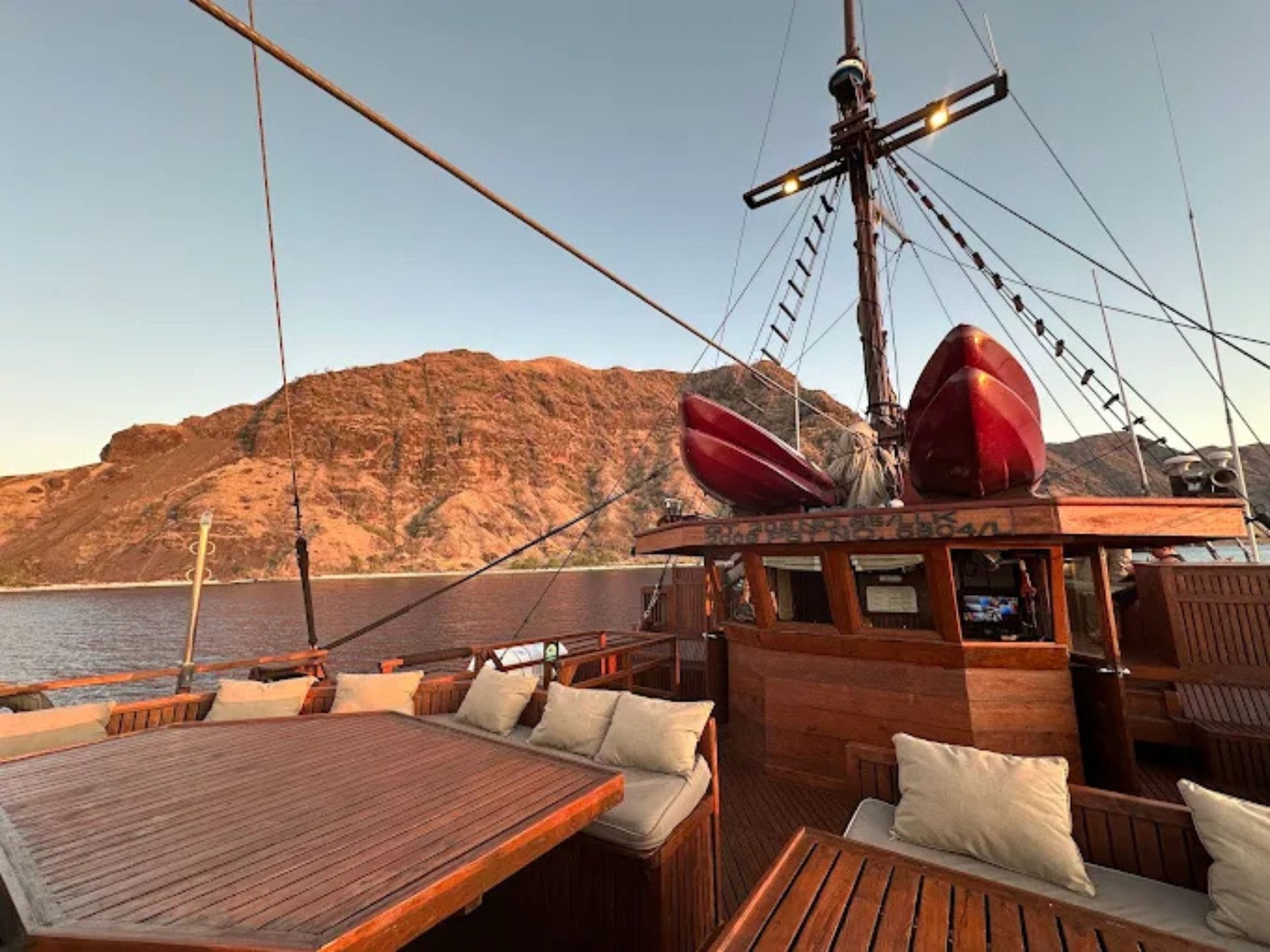 A deck view from a wooden boat at sunset, with seating areas, a mast, and two red kayaks near a mountainous coastline.