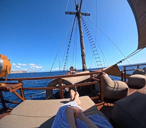 Feet resting on a deck chair aboard a wooden sailing boat on the deep blue sea under a clear sky.