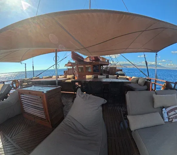 A spacious wooden deck on a yacht features lounge seating, a dining table, and a large canopy shade overlooking the sea.