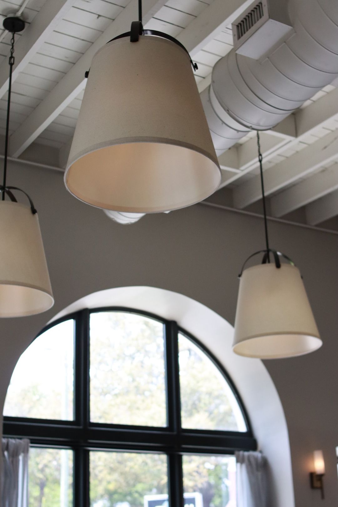 Three hanging beige pendant lights in a room with a large arched window and exposed ceiling pipes