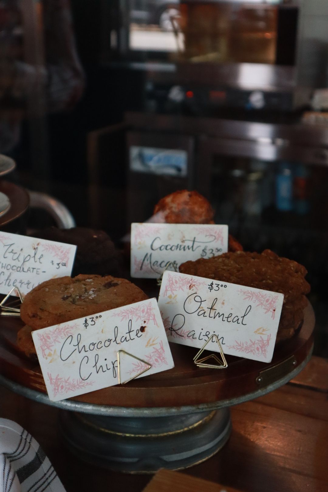 Dessert display with labeled baked goods on a glass pedestal in a café counter setting