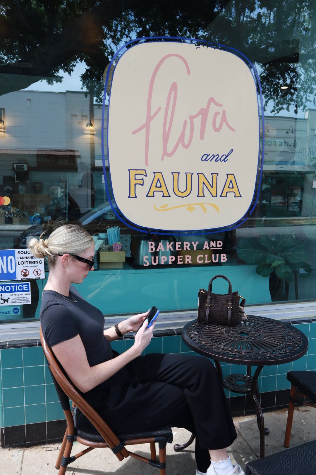 Woman sitting at a café table outside Flora and Fauna bakery and supper club, looking at a phone.