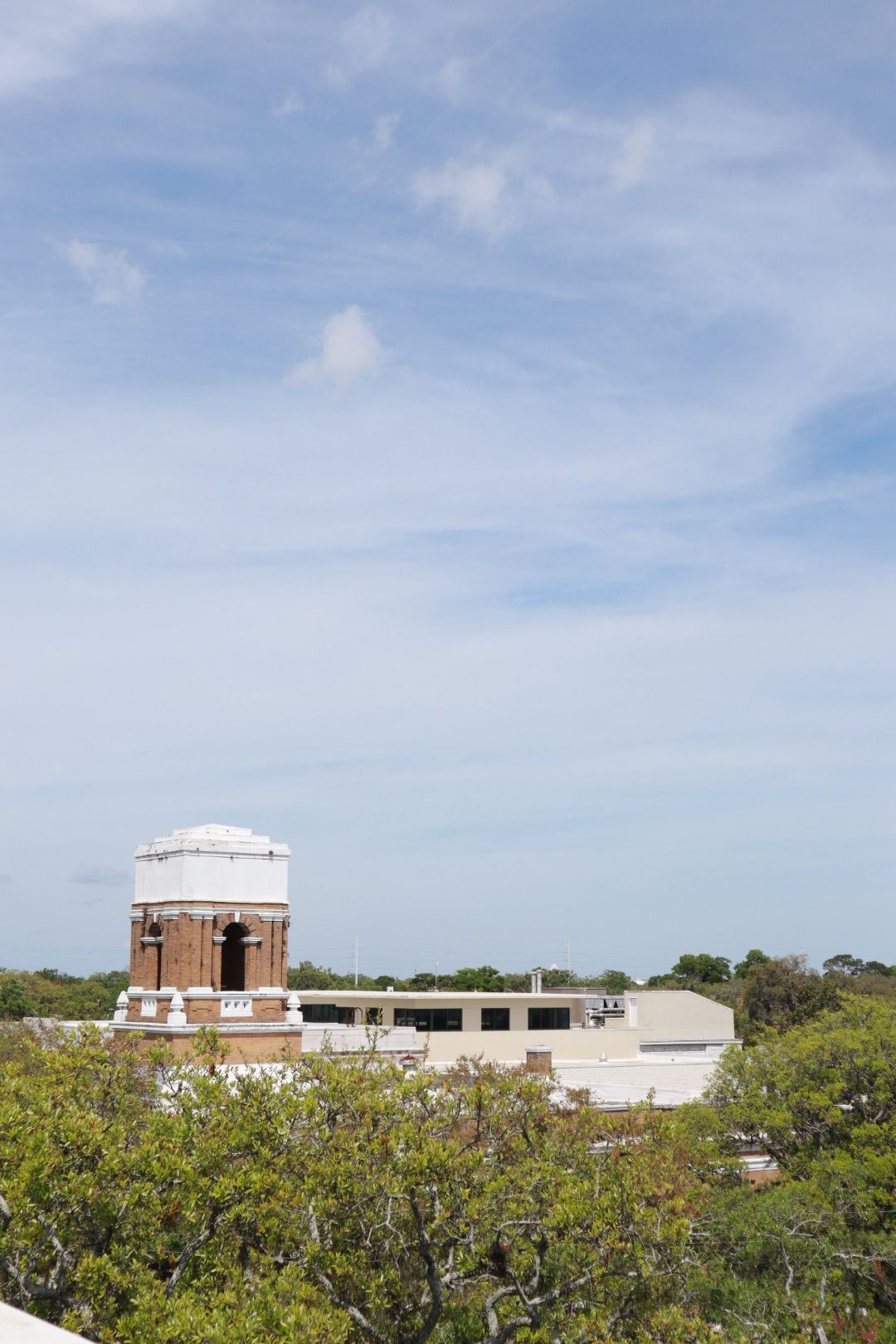 Distant brick water tower above low white buildings and green trees under a cloudy blue sky