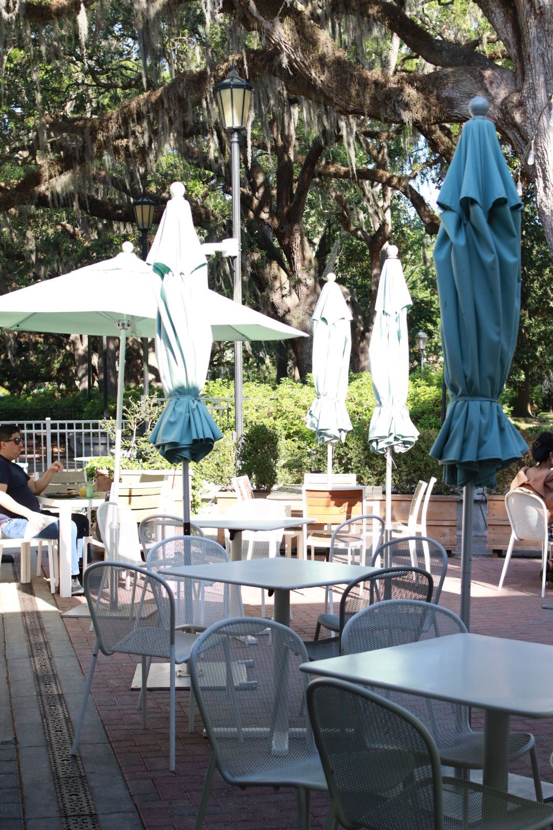 Outdoor café patio with tables, metal chairs, and closed white and blue umbrellas under trees.