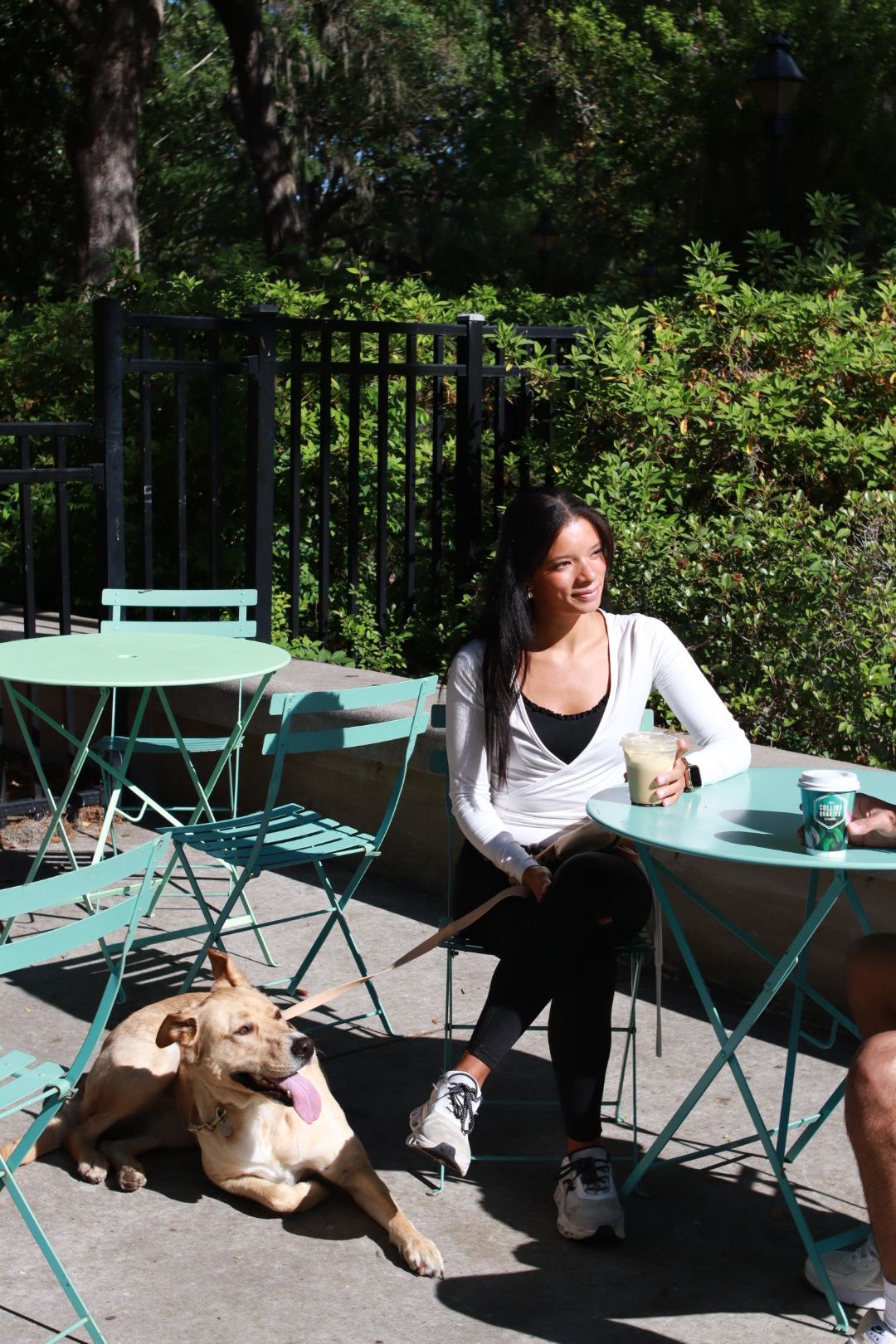 Woman sitting at a turquoise café table with two dogs in a sunny outdoor patio garden