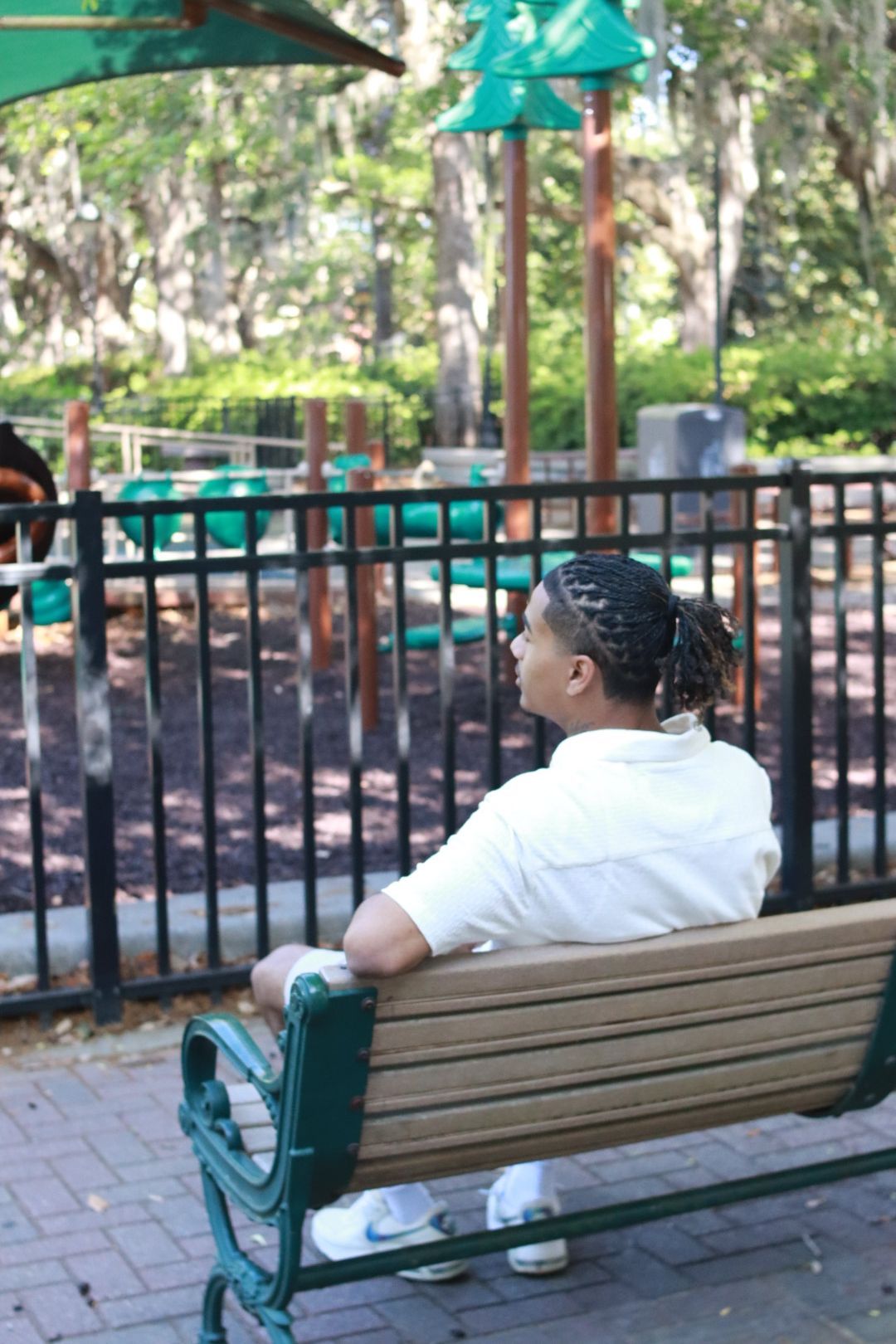 Person sitting on a park bench by a black fence, looking toward playground equipment in a wooded park.