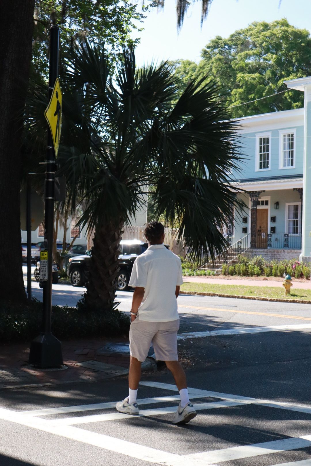 Person in white shirt and shorts crossing a street at a crosswalk near palm trees and houses.