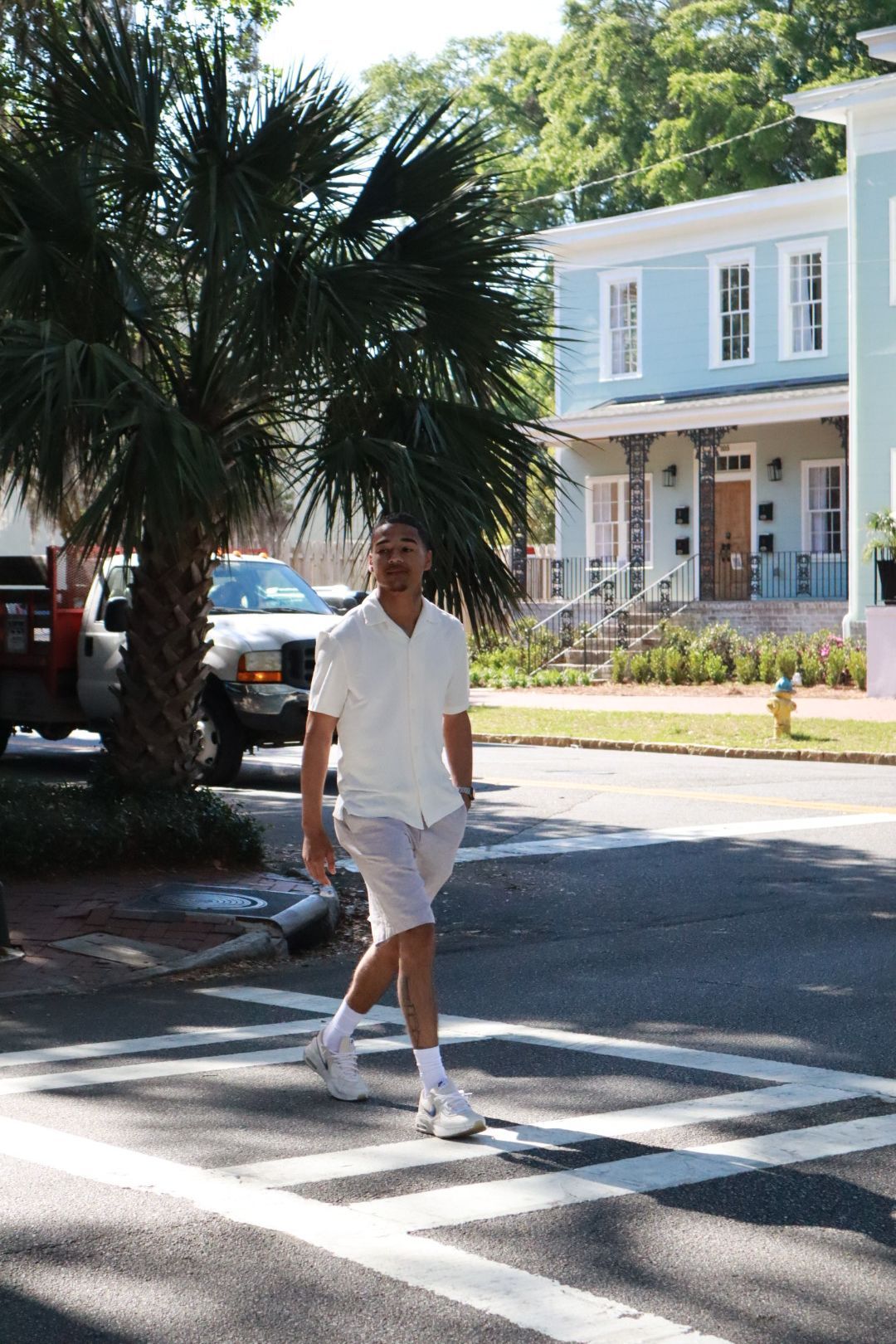Person in white walking across a sunlit street beside palm trees and pastel houses.