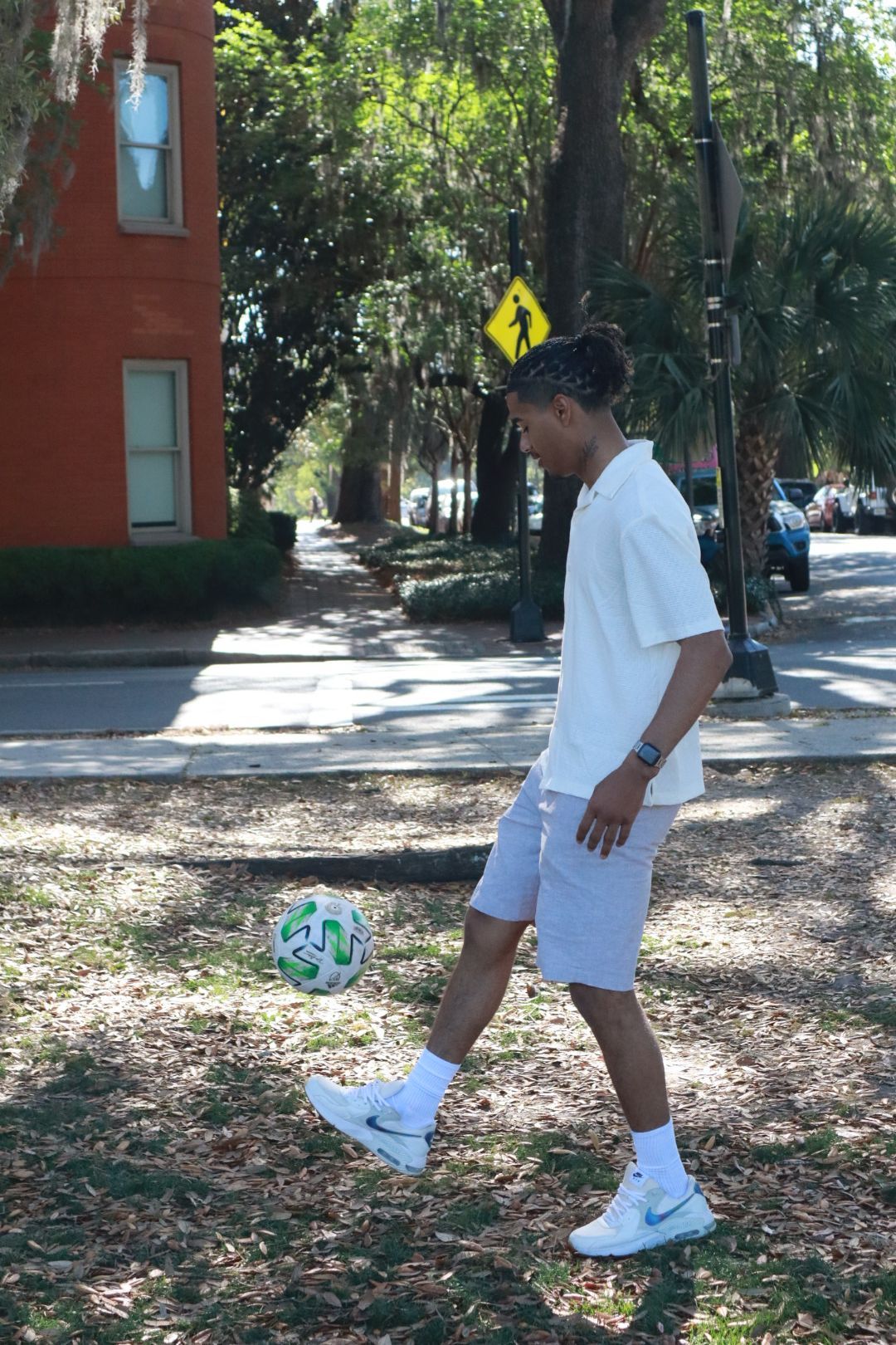Person in white athletic wear kicks a soccer ball on a shaded sidewalk near trees and a building.