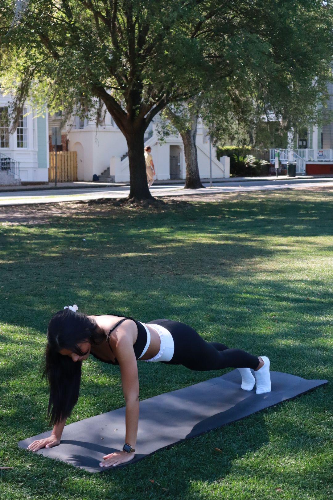 Person doing a plank on a yoga mat in a grassy park under a tree.