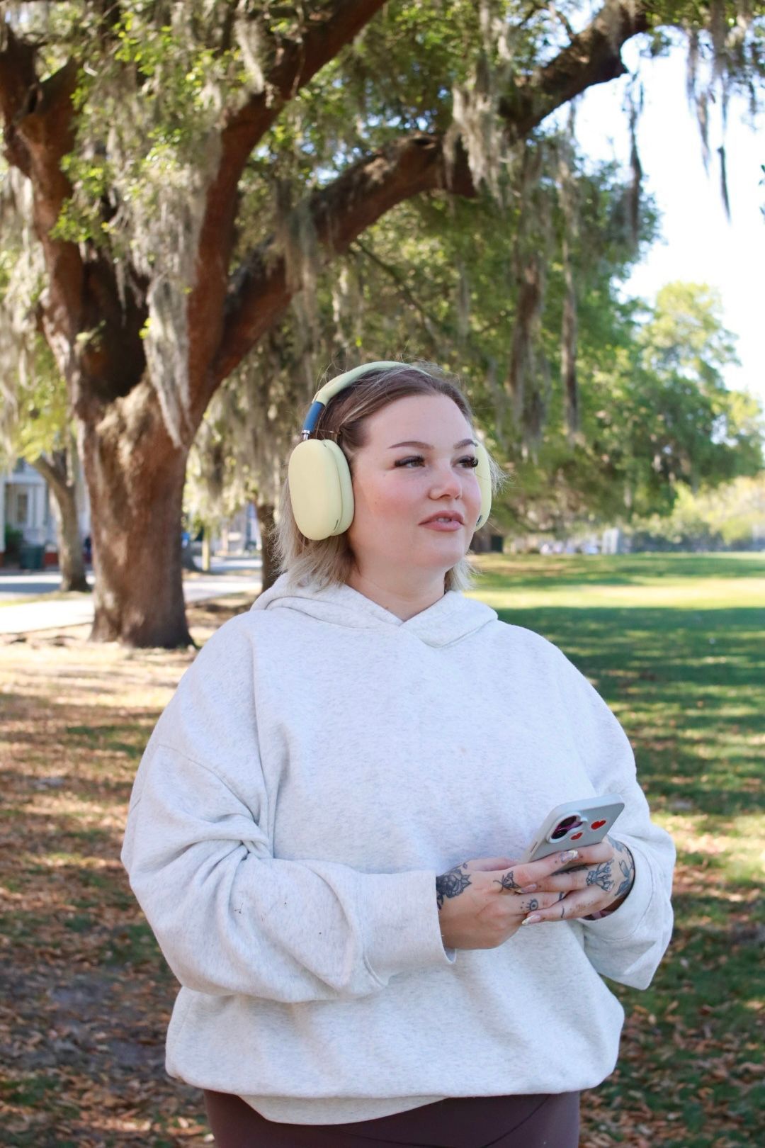 Person wearing headphones and a white hoodie stands outdoors holding a phone in a sunny park.