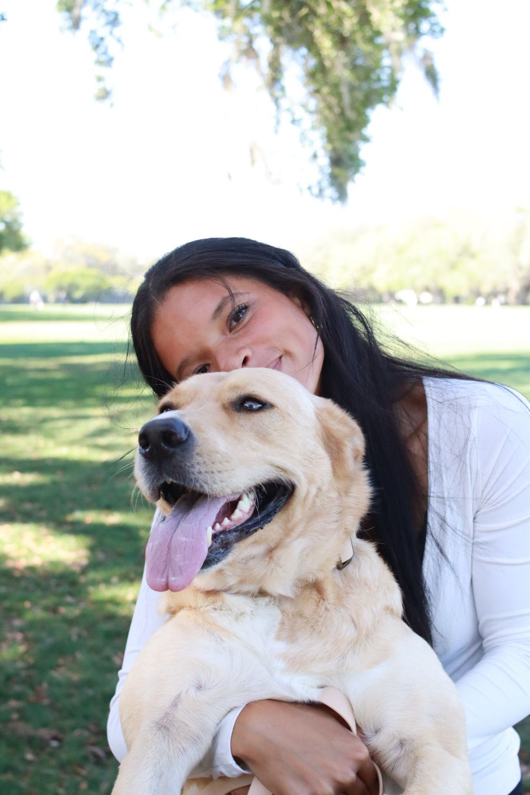 Woman hugging a golden retriever in a sunny park, both facing the camera and smiling