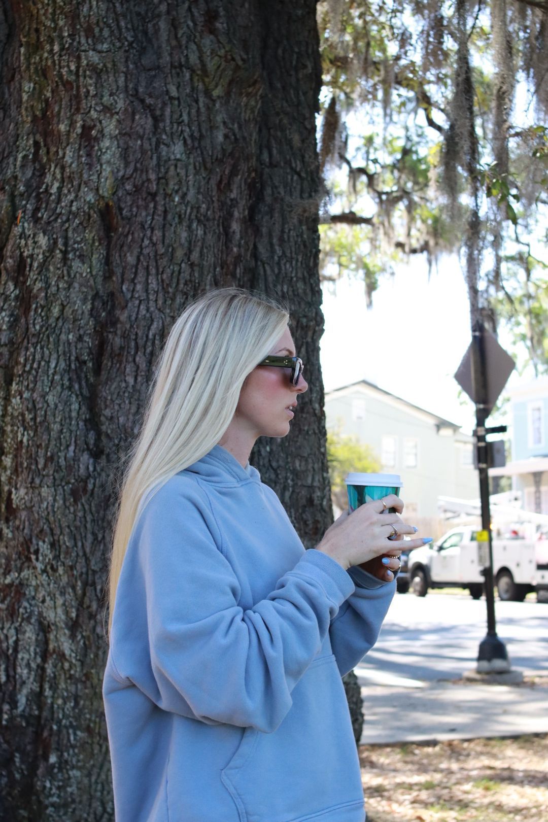 Person in a light blue sweater holding a cup beside a large tree on a sunny street