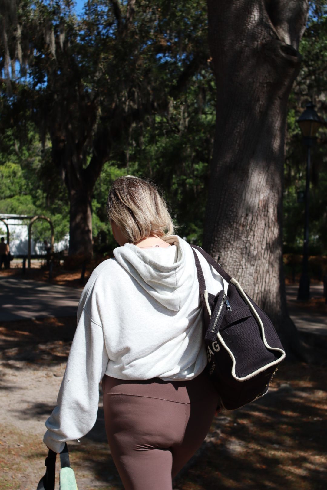 Person walking on a tree-lined path, wearing a white hoodie and carrying a black shoulder bag.