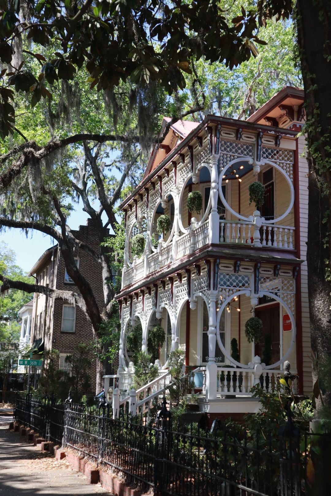 Victorian-style house with white trim and balconies beneath large oak trees on a sunny street
