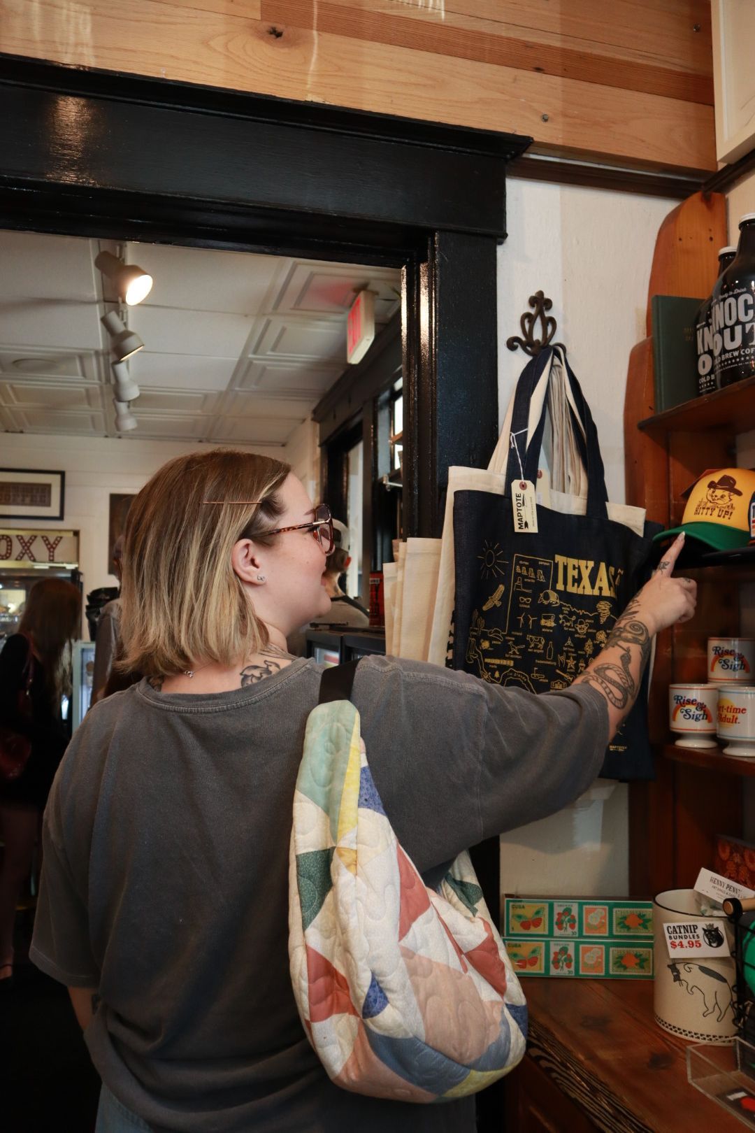 Woman reaching for a shelf in a shop, with a colorful tote bag over her shoulder
