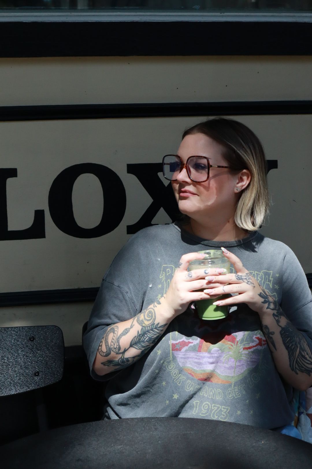 Person in glasses and gray shirt holding a drink, standing in front of a wall with large lettering.