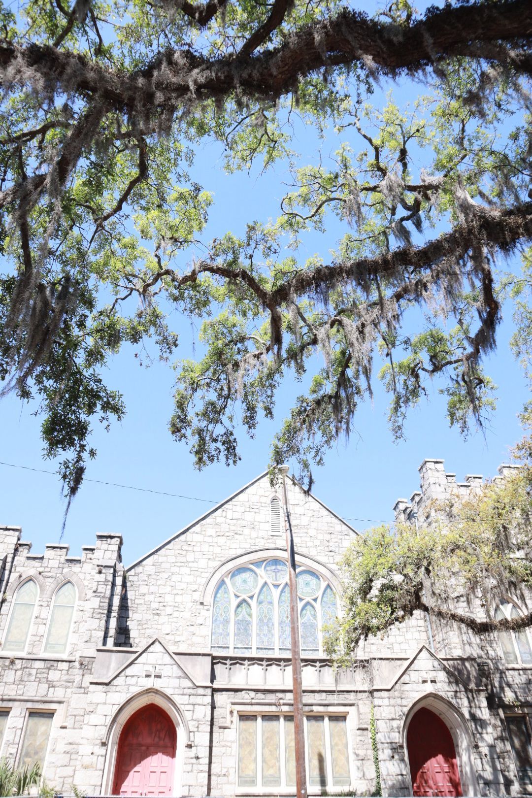 Stone church facade with red arched doors framed by hanging tree branches against a blue sky