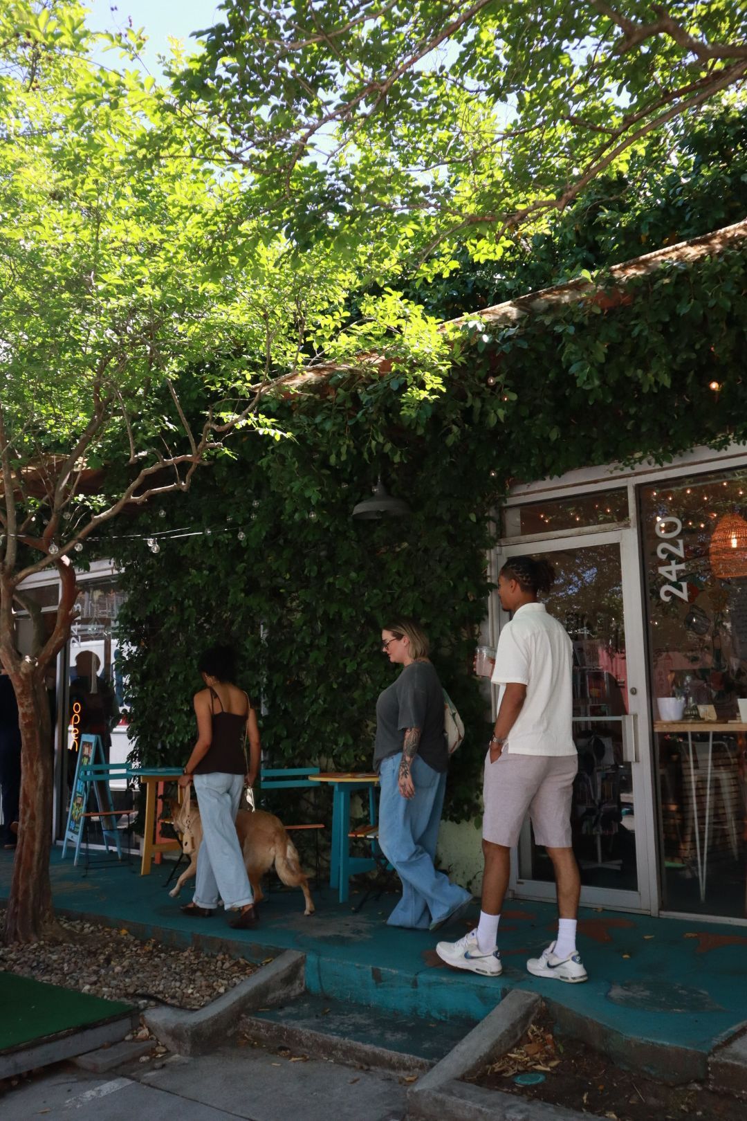 Three people walking past a green café patio under trees, with turquoise tables and a storefront entrance.