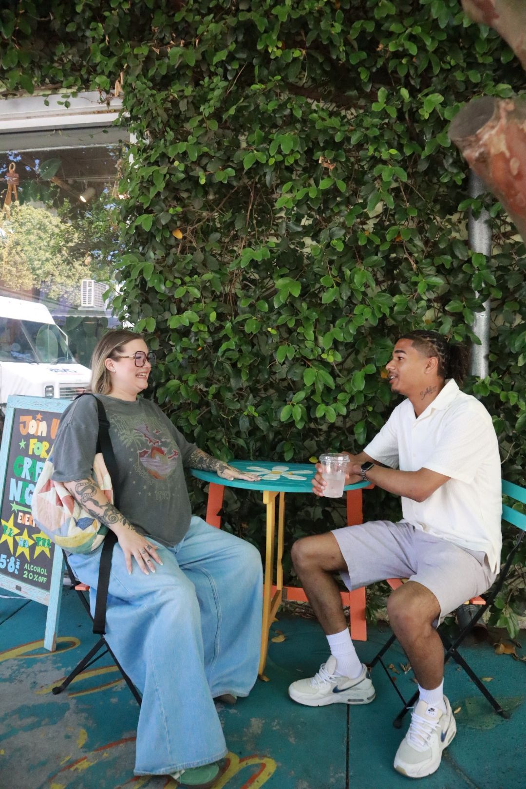 Two people sitting and talking at a small outdoor table beside a leafy wall