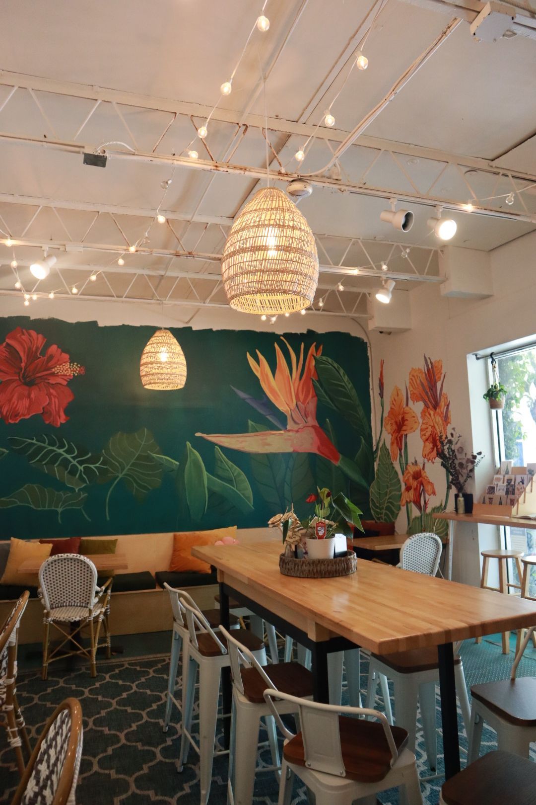 Bright café interior with wooden tables, woven pendant lights, and a green floral mural on the wall
