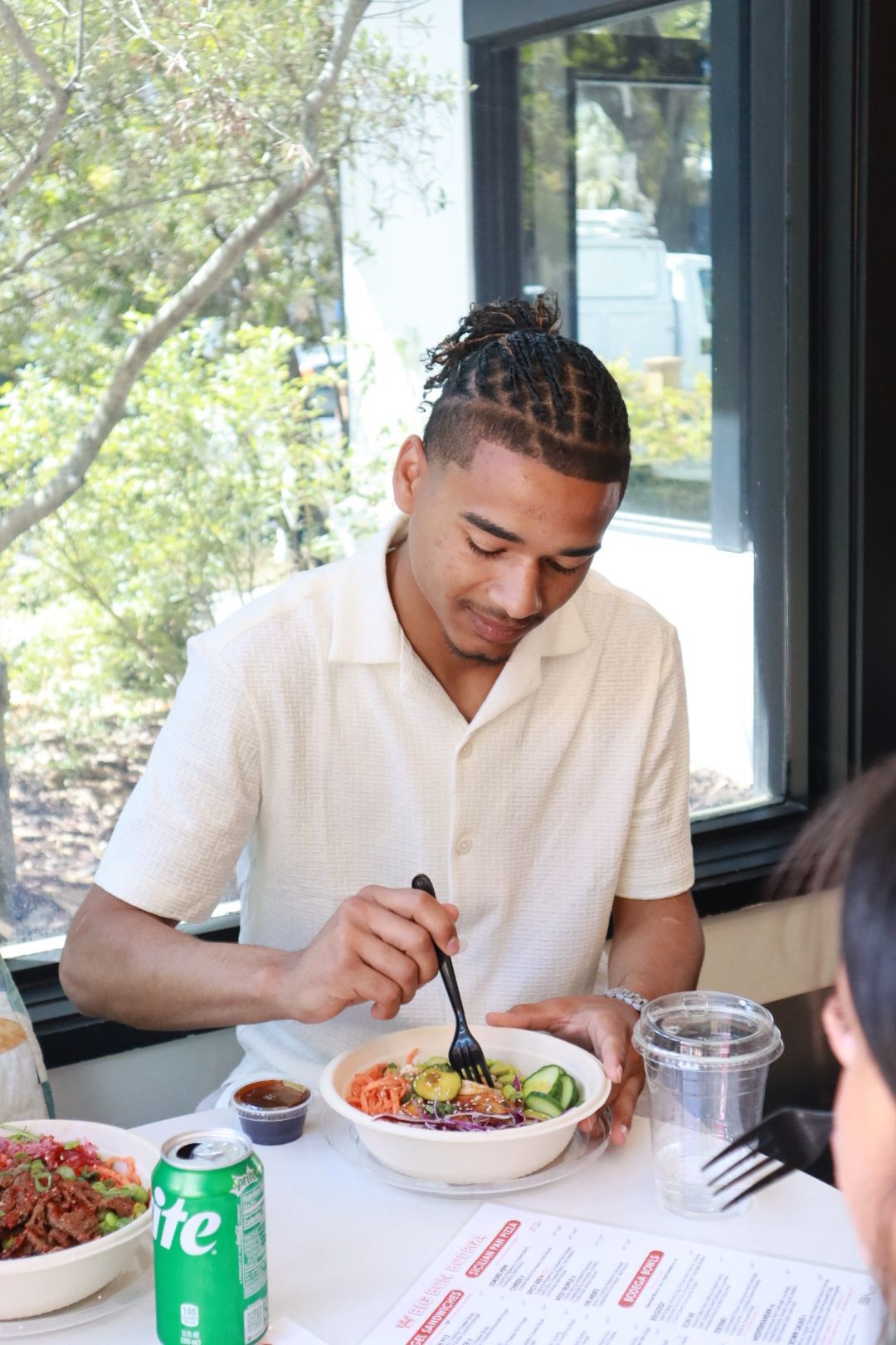 Man eating a salad at a restaurant table by a window, with a soda can nearby.