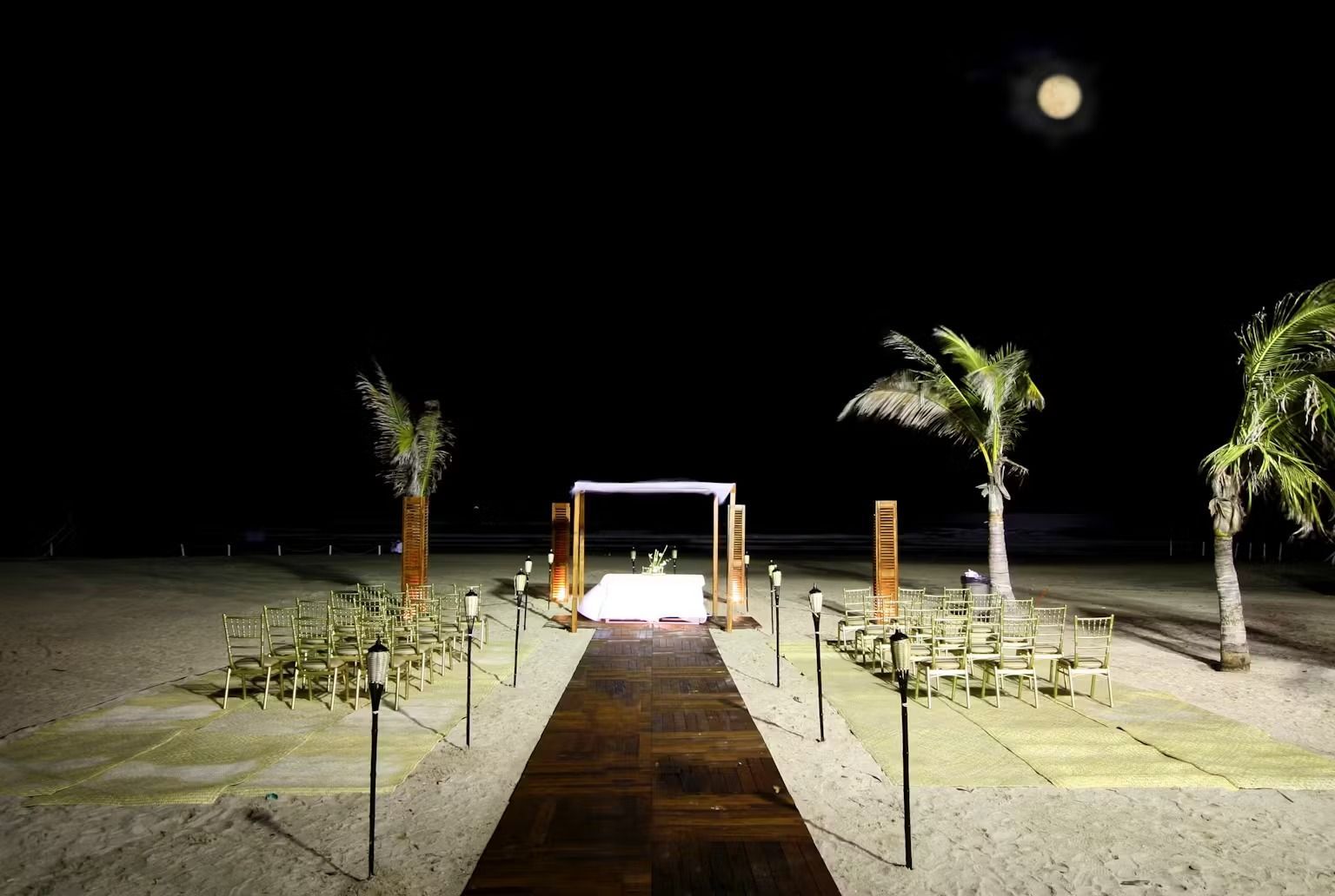 A wooden walkway leading to a wedding ceremony on a beach at night.