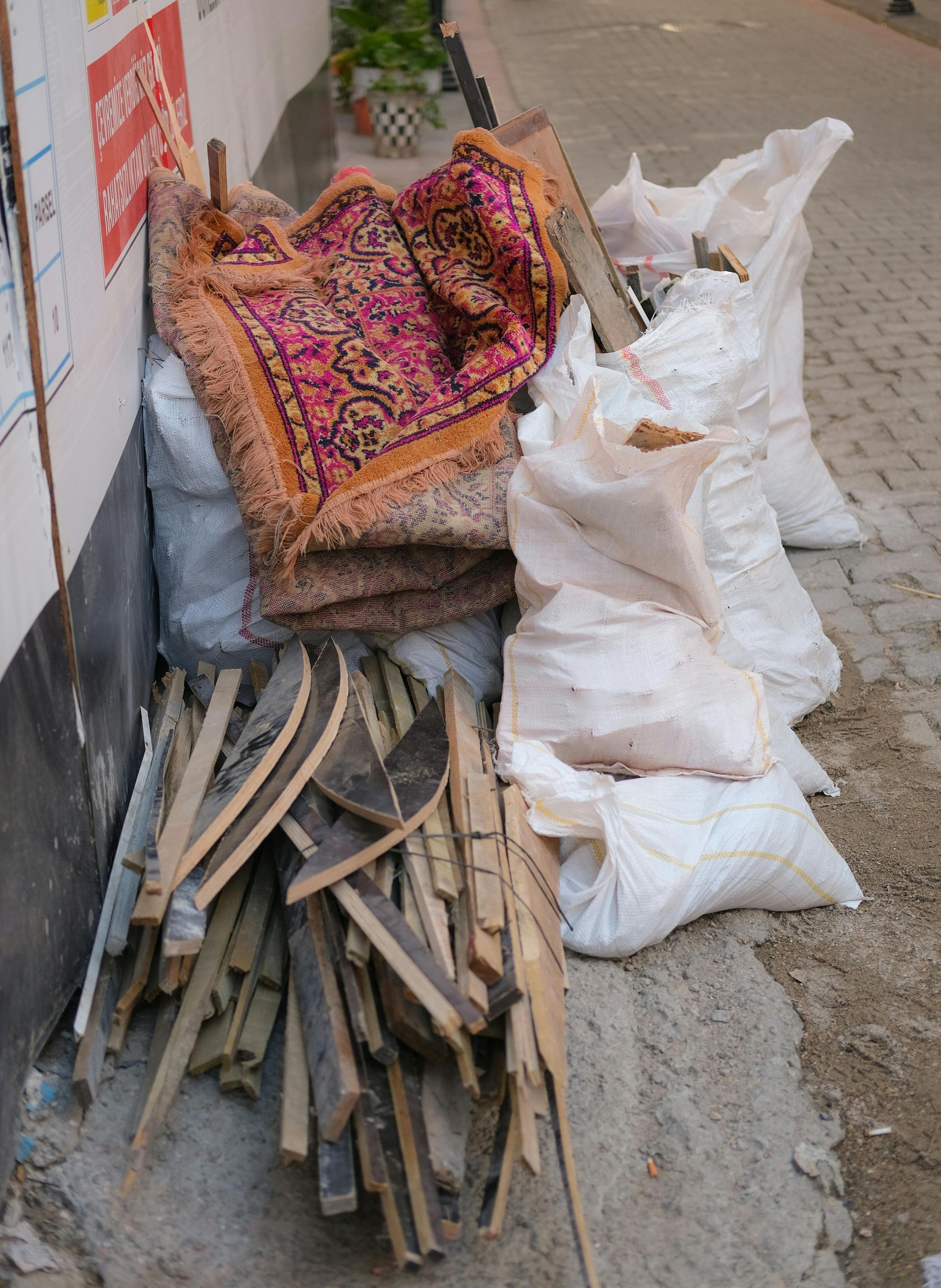 Stacked firewood and white sacks beside a wall on a sidewalk, with a colorful blanket draped over the pile.