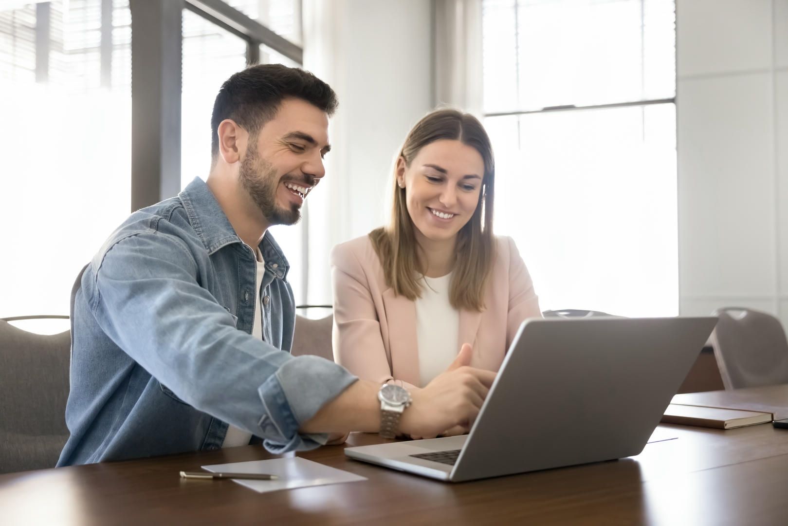 Man and woman smiling, looking at laptop screen together in a well-lit office setting.