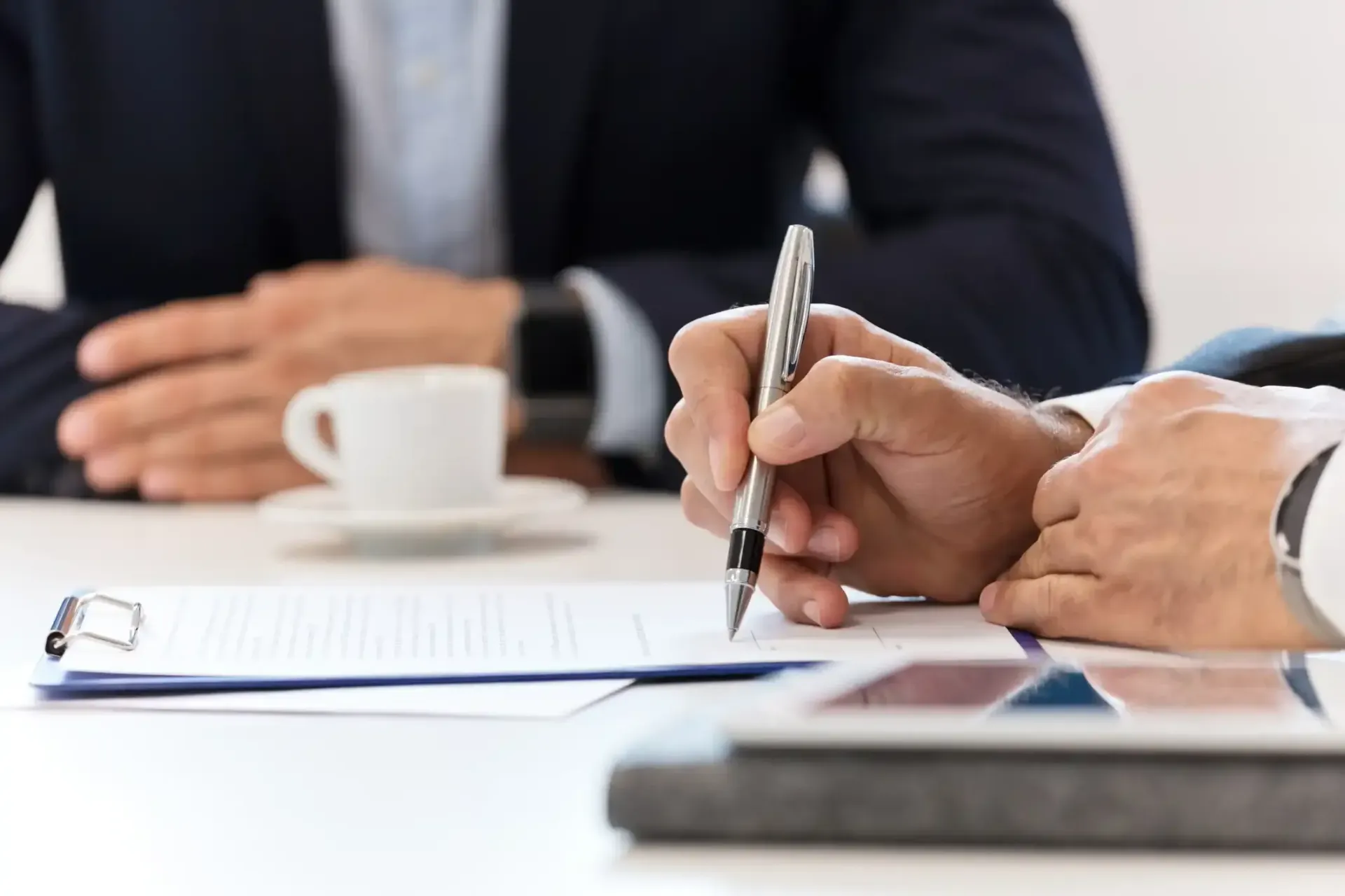Person signing document with another person in the background, table, pen, coffee cup, and tablet.