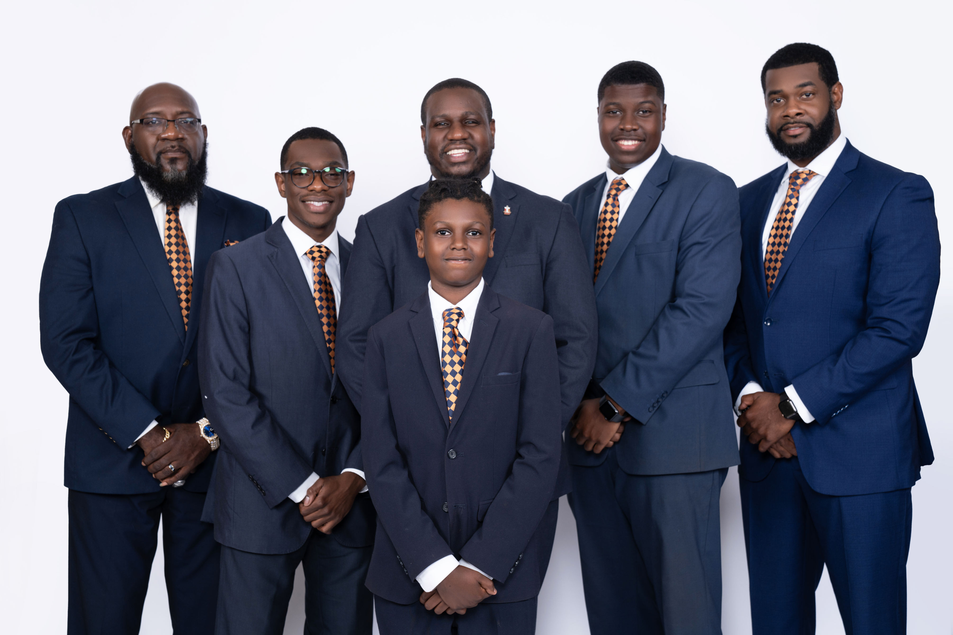 Six men in suits, smiling, posing together against a white background; all wearing gold patterned ties.