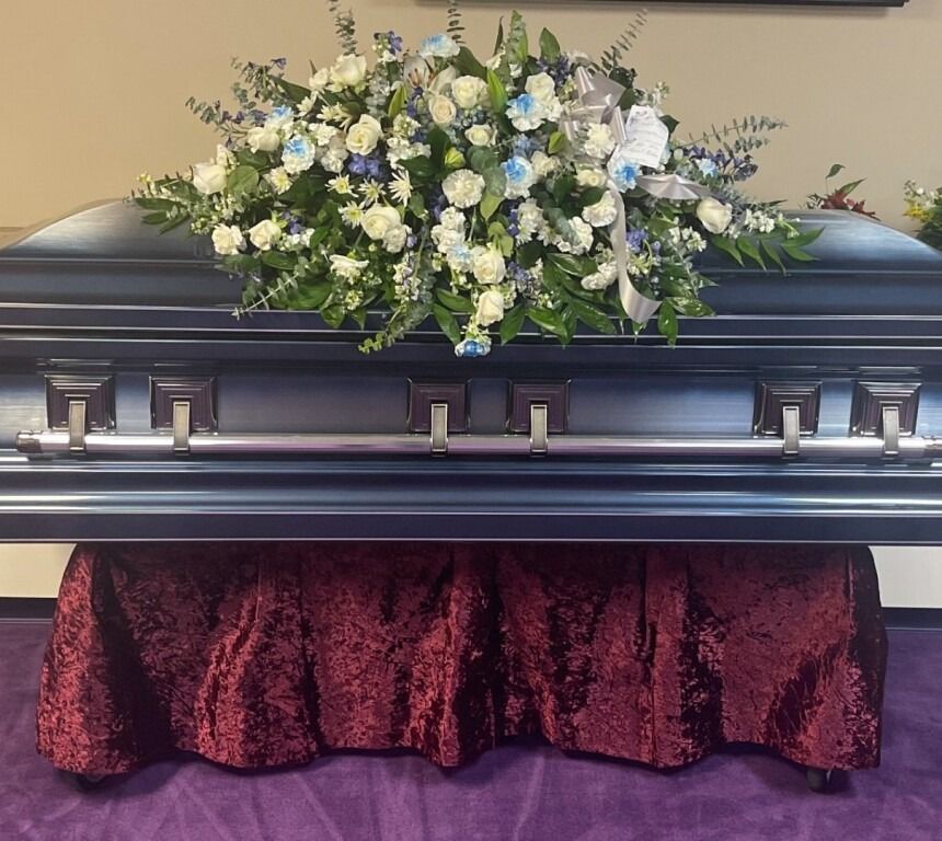 Blue casket with a floral arrangement, draped with a burgundy cloth on a purple surface.