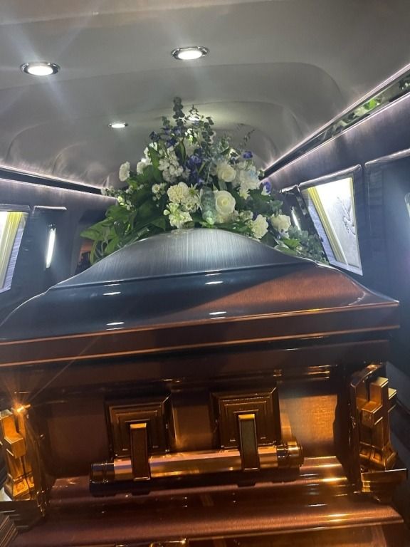 Brown casket inside a hearse, adorned with a white and blue floral arrangement.
