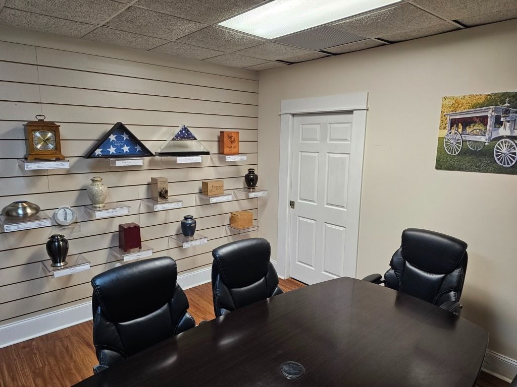 Meeting room with a display of urns and flags on shelves; a closed door and three chairs around a table are in the room.