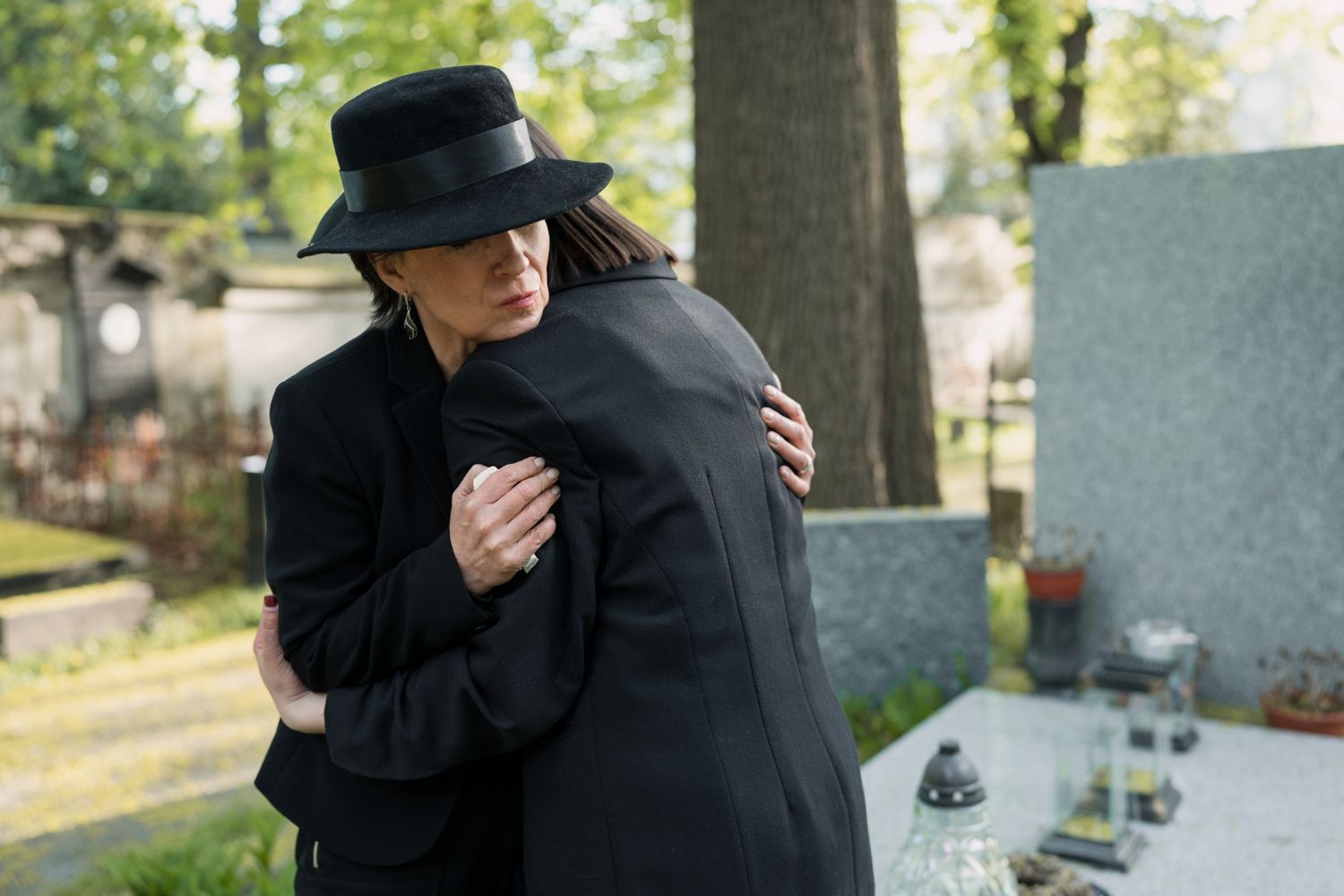 Two people in black clothing embrace at a cemetery near a headstone.