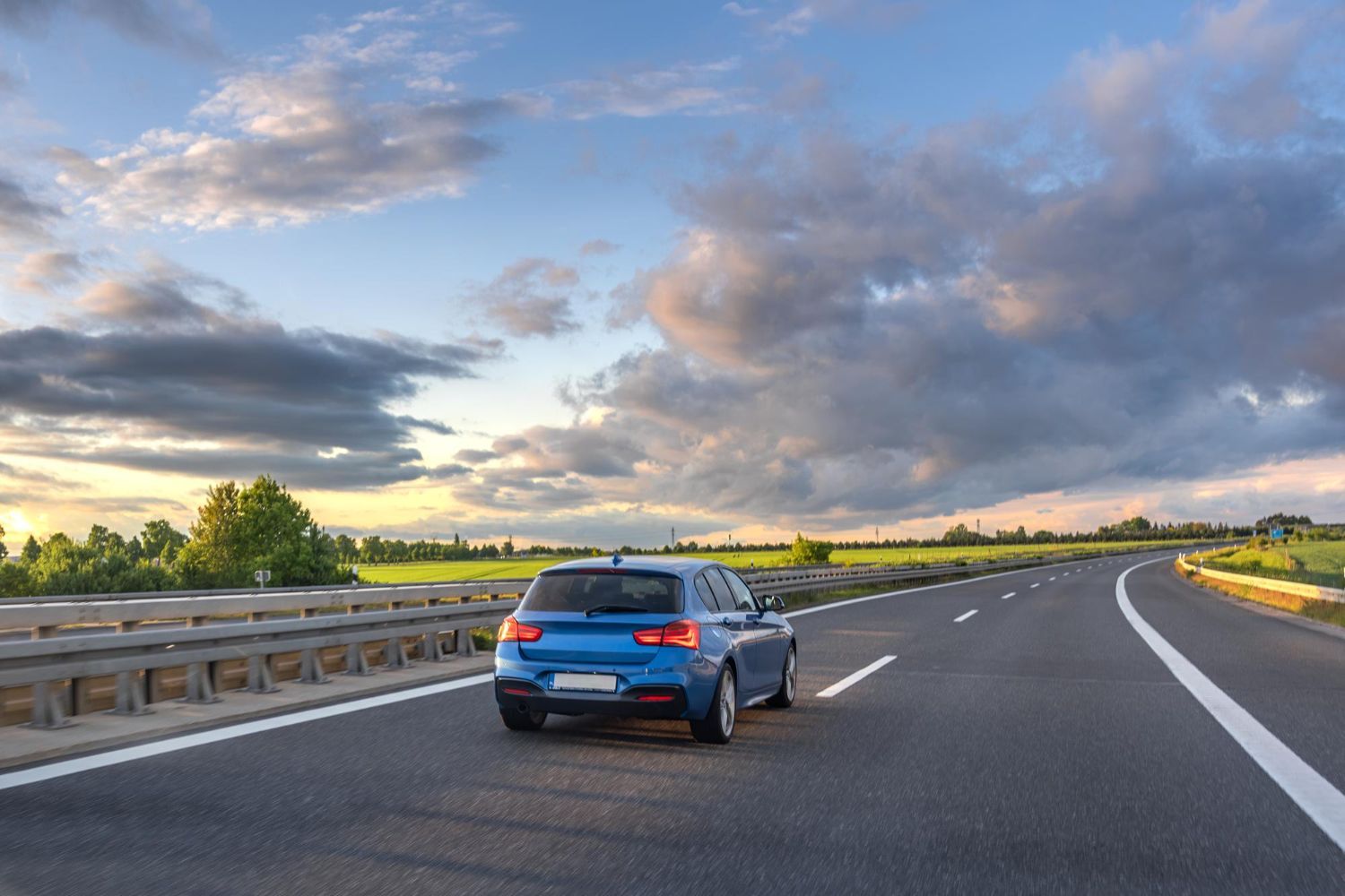 Blue car driving on a highway with cloudy sky and green fields.