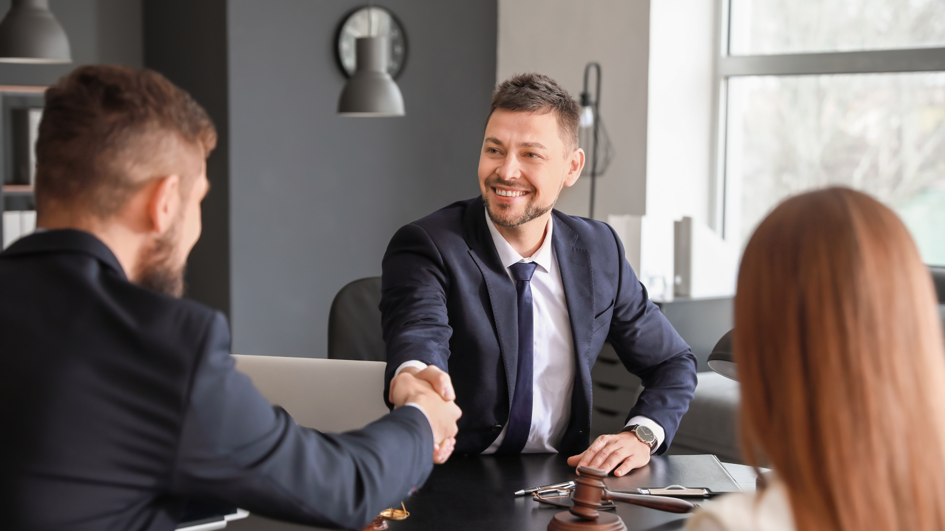 Man in suit shakes hands with another man, smiling. A woman is seated nearby.
